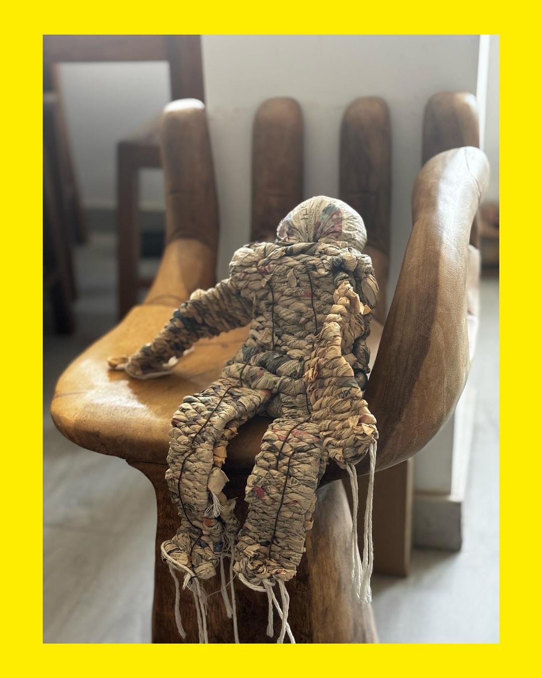 Foto de una figura humana, semejante a una marioneta, hecha con pedazos de periódicos torcidos y cuerdas de nylon. Está sentada sobre un asiento escultórico de madera en forma de mano.